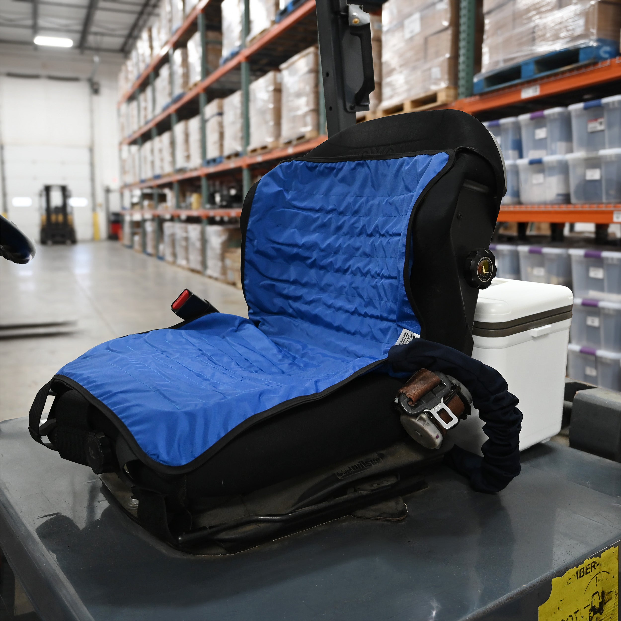 A blue and black circulating cooling seat attached to a 13-quart cooling reservoir installed on a vehicle seat, resting on a forklift platform in a warehouse setting.