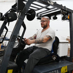 A man with tattoos and glasses sits in the driver's seat of a Toyota forklift inside a warehouse, wearing a white t-shirt and dark gray sweatpants. He is sitting on a blue circulating cooling seat attached to a 30-quart cooling reservoir. 