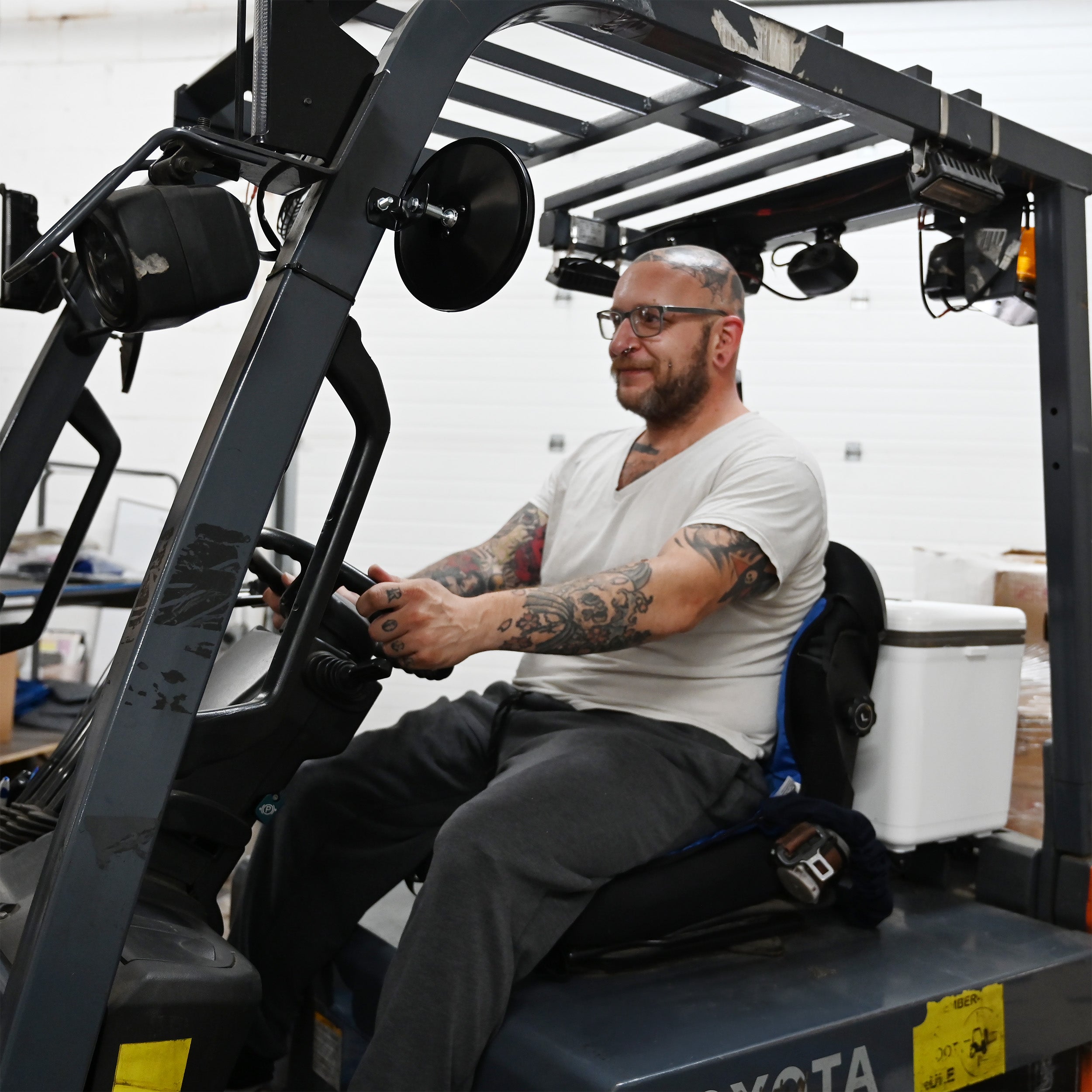 A man with tattoos and glasses sits in the driver's seat of a Toyota forklift inside a warehouse, wearing a white t-shirt and dark gray sweatpants. He is sitting on a blue circulating cooling seat attached to a 30-quart cooling reservoir. 