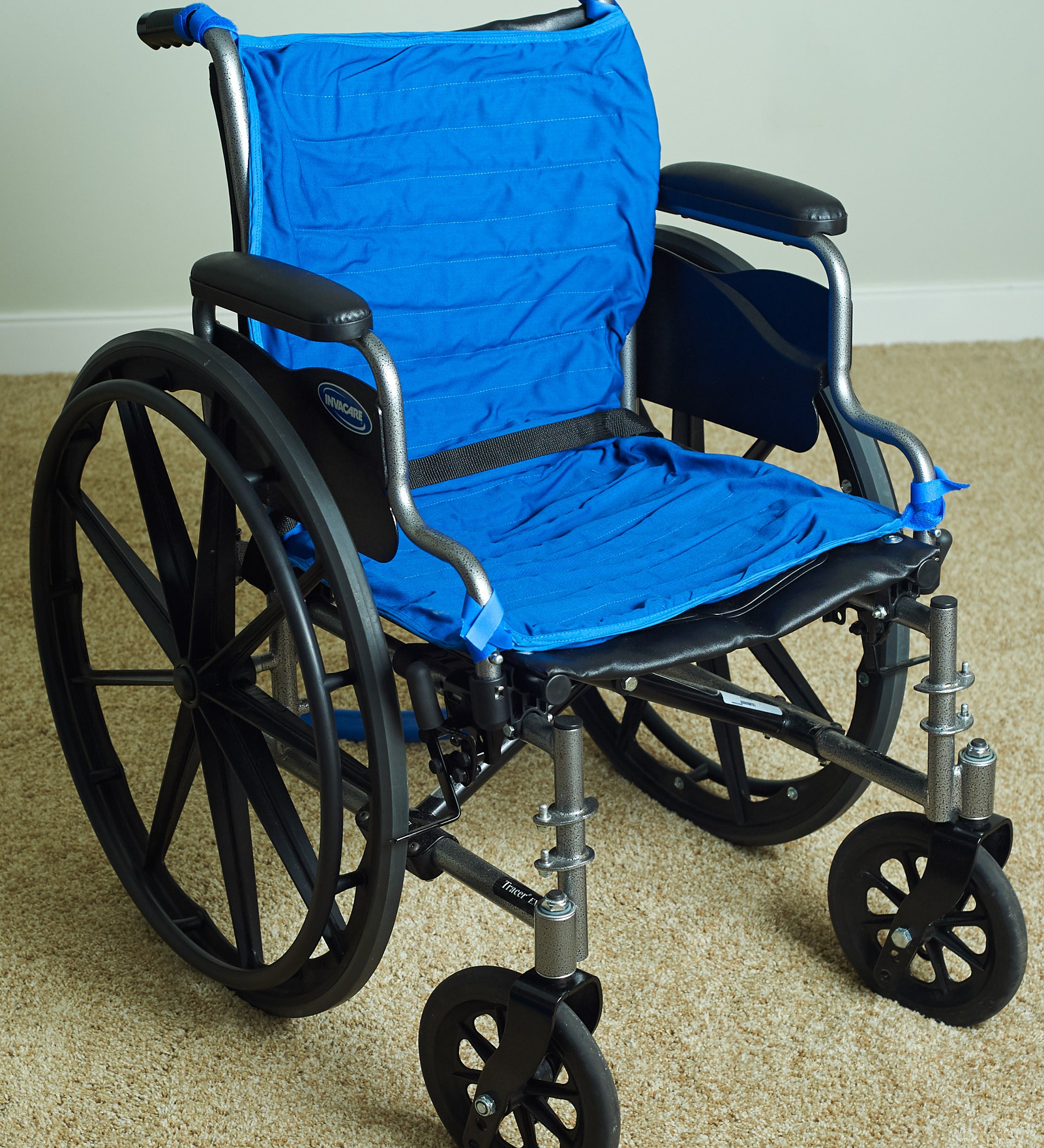 A manual wheelchair with a bright blue circulating cooling water seat and backrest, black wheels, and a silver metal frame, positioned on a beige carpeted floor against a light wall.