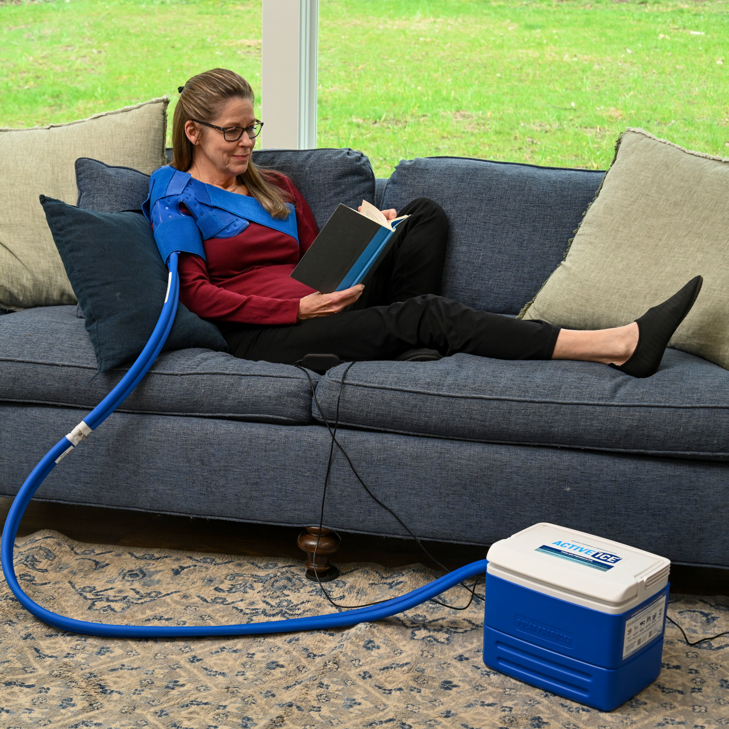 A woman relaxes on a blue couch reading a book while wearing a blue broad shoulder therapy pad. A blue hose connects her to a blue and white portable cooling Active Ice® unit on the floor in a living room setting.