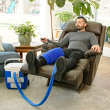 A man reclining in a brown leather armchair with his legs extended, wearing a blue large full circumference knee therapy pad. A blue and white 16-quart Active Ice® Cold Therapy system connected by a blue hose is placed on the floor beside him in a bright living room.