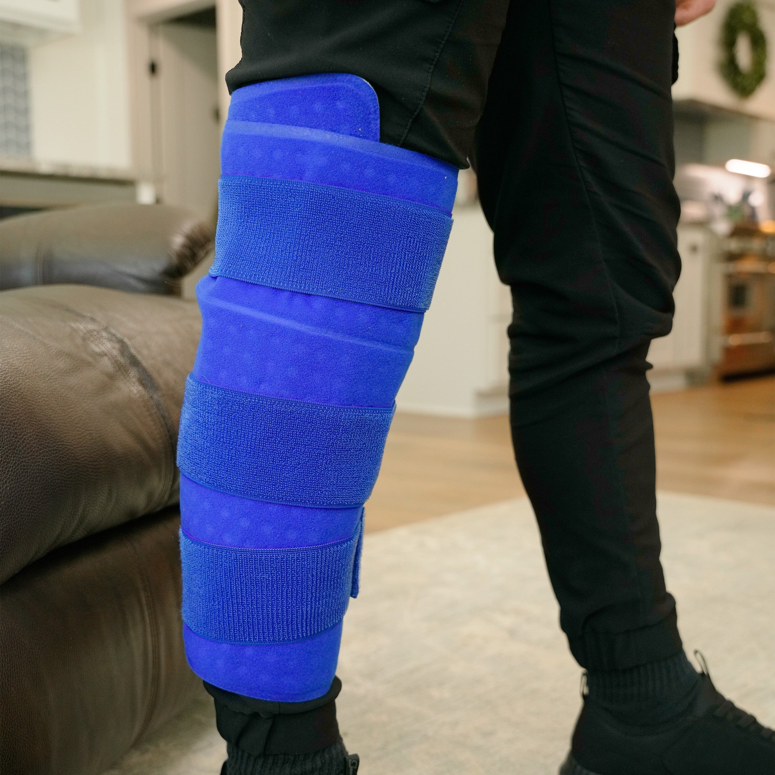 A person wearing a bright blue Large Full Circumference knee therapy pad on their right leg, standing in a living room with a couch and kitchen visible in the background.