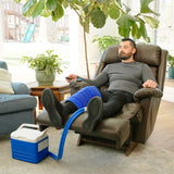A man reclining in a brown leather armchair with his legs extended, wearing a blue large full circumference knee therapy pad. A blue and white 9-quart Active Ice® Cold Therapy system connected by a blue hose is placed on the floor beside him in a bright living room.