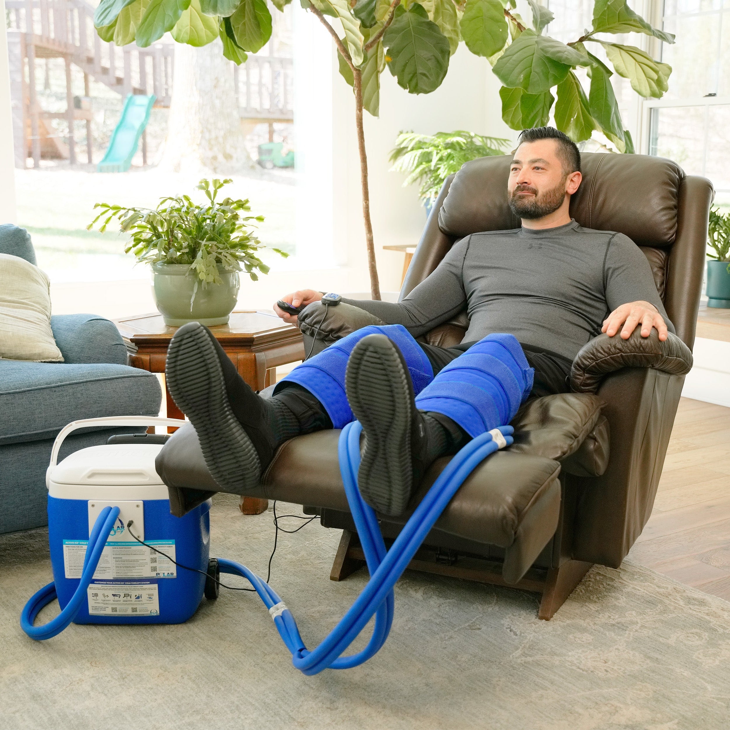 A man reclining in a brown leather armchair with his legs extended, wearing a double blue large full circumference knee therapy pad. A blue and white 16-quart Active Ice® Cold Therapy system connected by a blue hose is placed on the floor beside him in a bright living room.