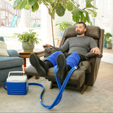 A man reclining in a brown leather armchair with his legs extended, wearing a double blue large full circumference knee therapy pad. A blue and white 9-quart Active Ice® Cold Therapy system connected by a blue hose is placed on the floor beside him in a bright living room.