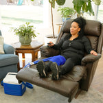 A woman reclining in a brown leather armchair with her legs extended, wearing a blue small full circumference knee therapy pad. A blue and white 9-quart Active Ice® Cold Therapy system connected by a blue hose is placed on the floor beside her in a bright living room.