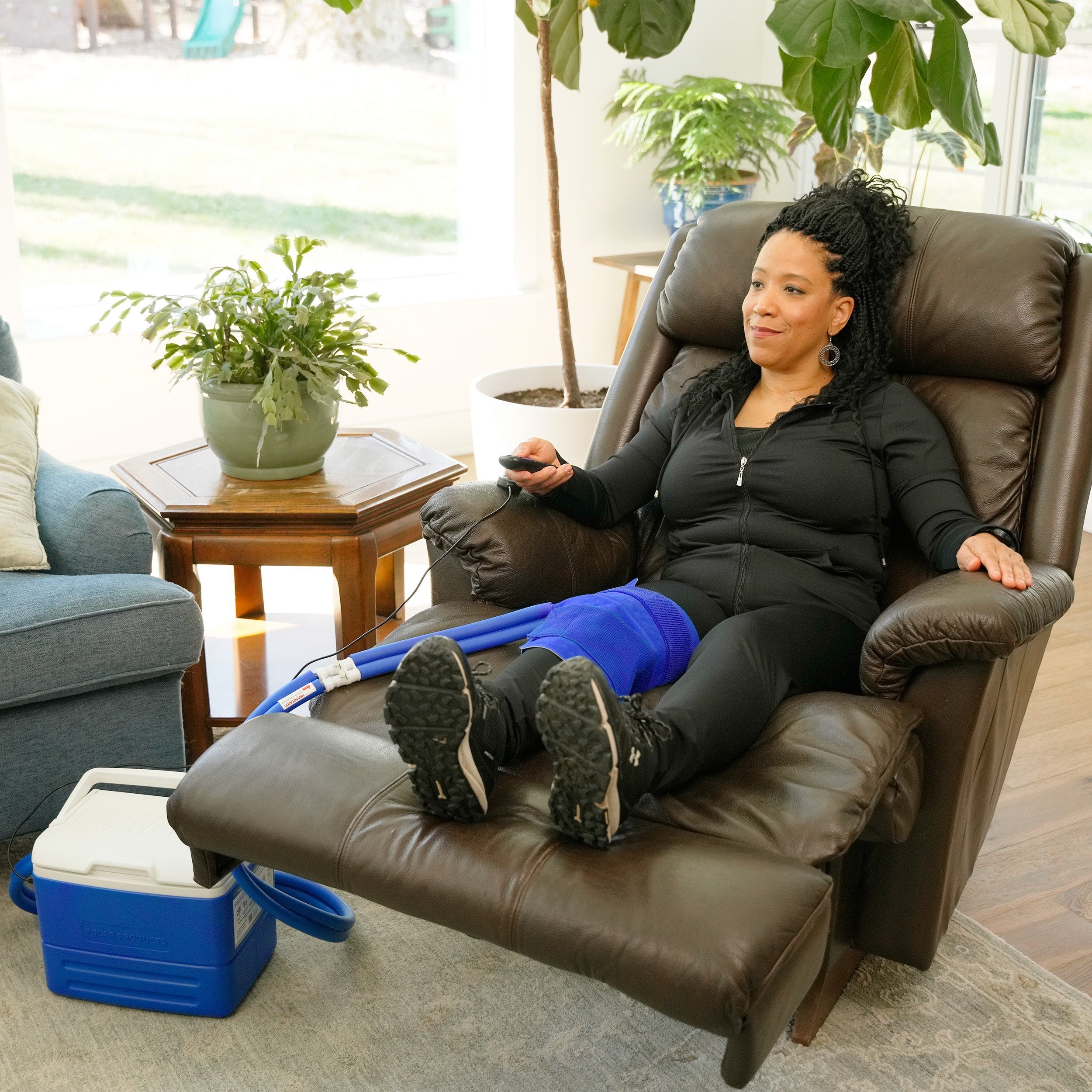 A woman reclining in a brown leather armchair with her legs extended, wearing a blue small full circumference knee therapy pad. A blue and white 9-quart Active Ice® Cold Therapy system connected by a blue hose is placed on the floor beside her in a bright living room.