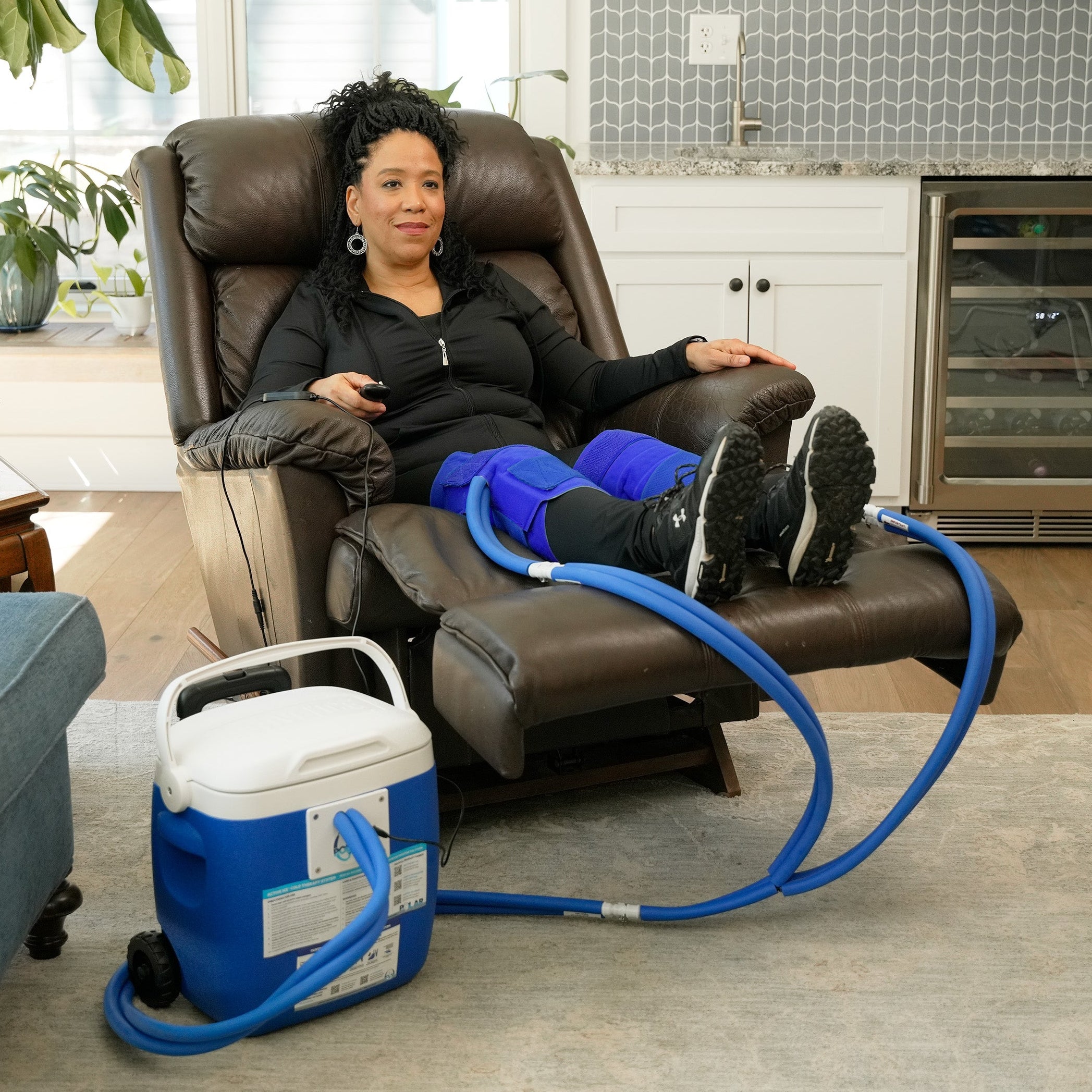 A woman reclining in a brown leather armchair with her legs extended, wearing a double blue small full circumference knee therapy pad. A blue and white 16-quart Active Ice® Cold Therapy system connected by a blue hose is placed on the floor beside her in a bright living room.