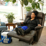 A woman reclining in a brown leather armchair with her legs extended, wearing a blue small full circumference knee therapy pad. A blue and white 16-quart Active Ice® Cold Therapy system connected by a blue hose is placed on the floor beside her in a bright living room.