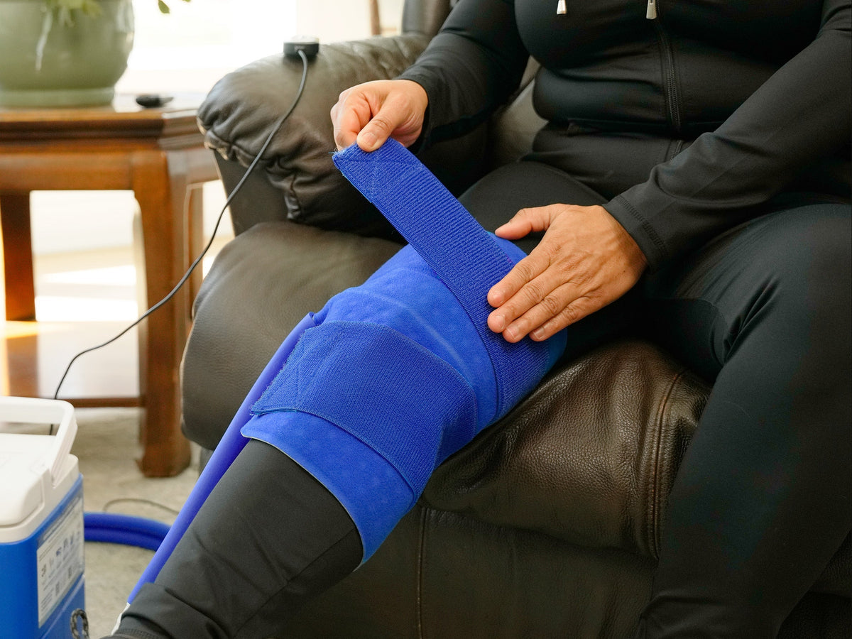 Woman applying a blue, small full circumference knee therapy pad on her right knee while sitting on a brown leather chair in a bright living room.