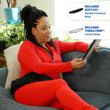 A woman sits on a gray couch wearing a bright red matching outfit and holding a tablet, with two callout graphics on the right showing included accessories: a black Soft Ice universal wrap and a gray Thera-Temp cervical wrap.
