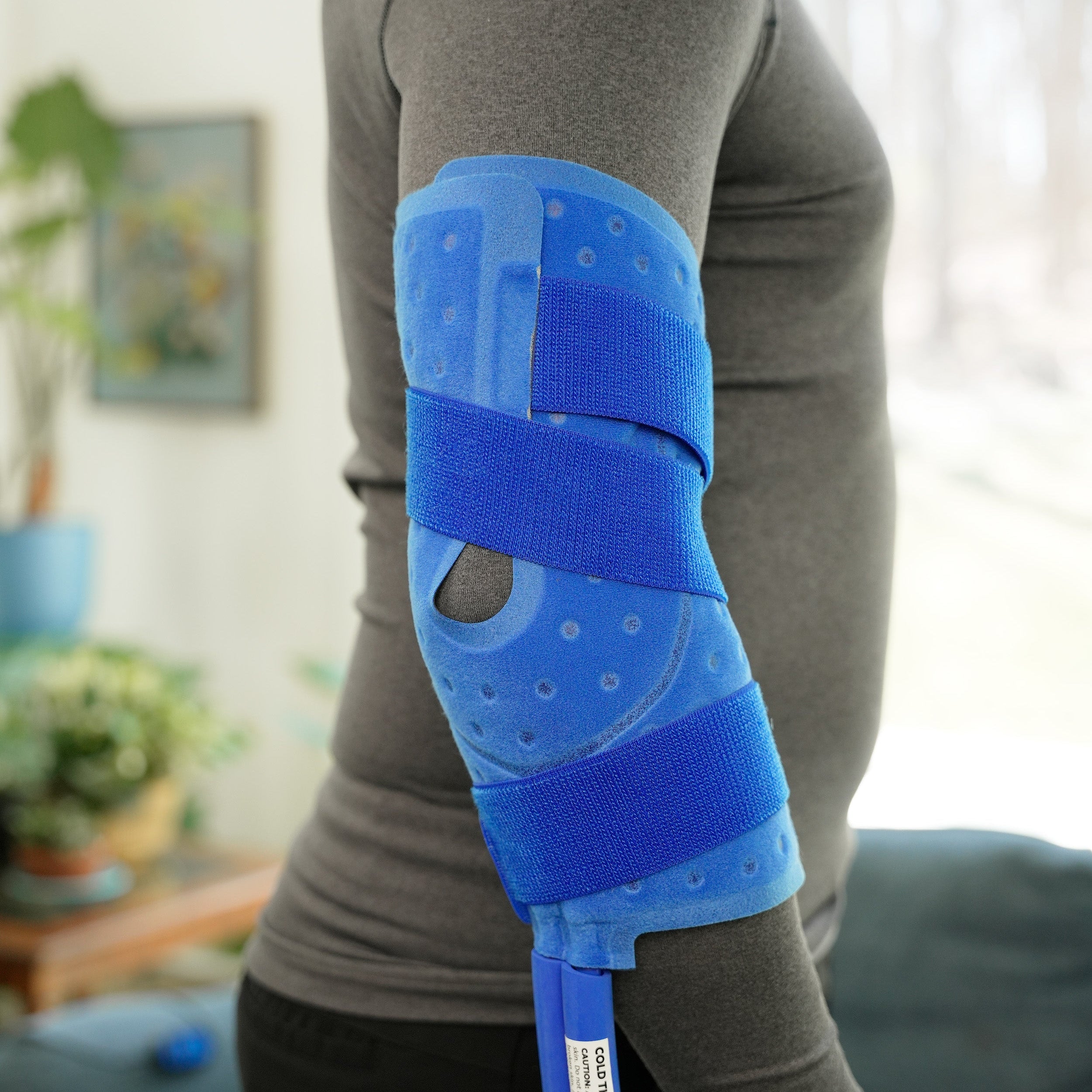 A person wearing a blue knee & joint polar active ice® cold therapy pad on their elbow, in a home setting with a couch and plants in the background.