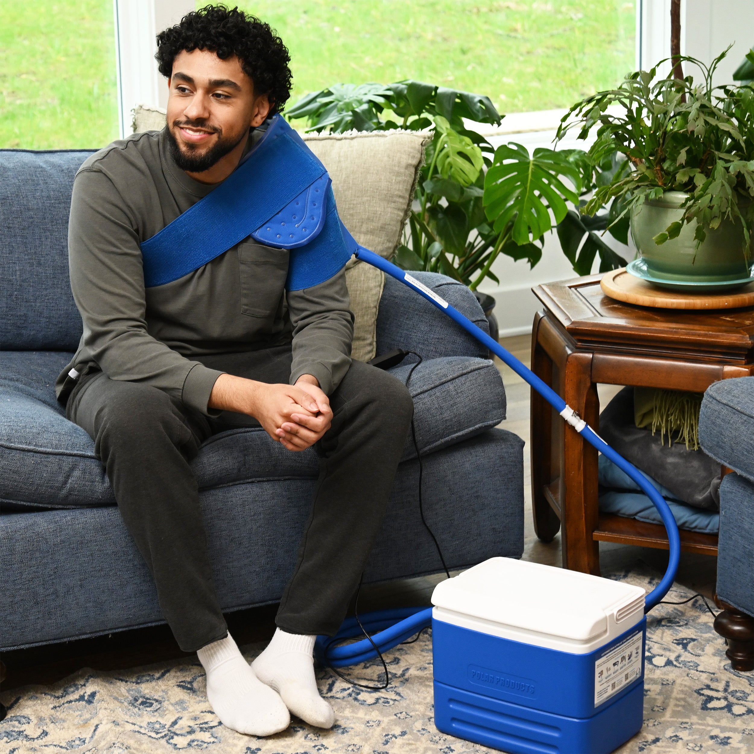 Man sitting on a couch using a blue Active Ice® Broad Shoulder & Upper Arm cold therapy system in a living room.