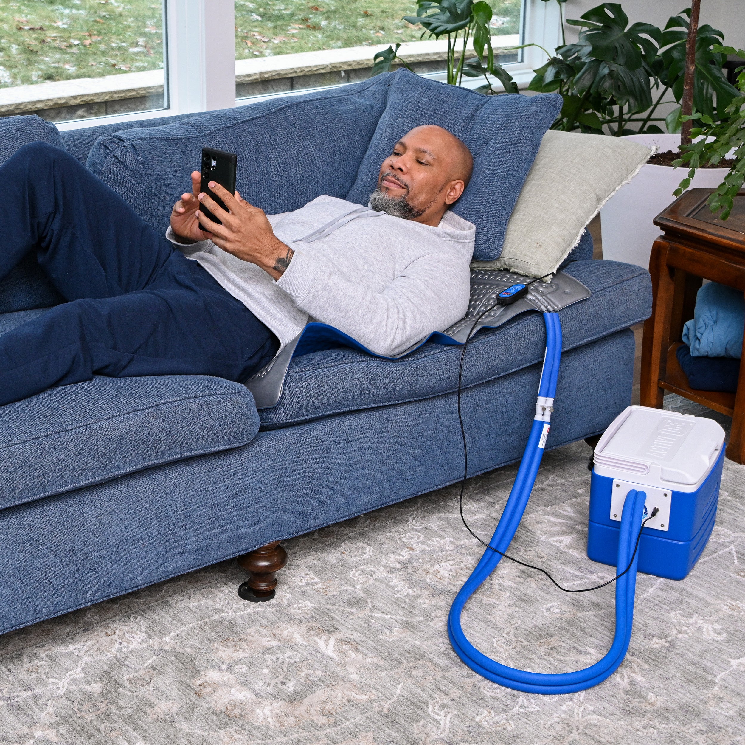 Man sitting on a blue couch using an active ice® cold therapy system while laying on the blanket therapy pad, using his phone.
