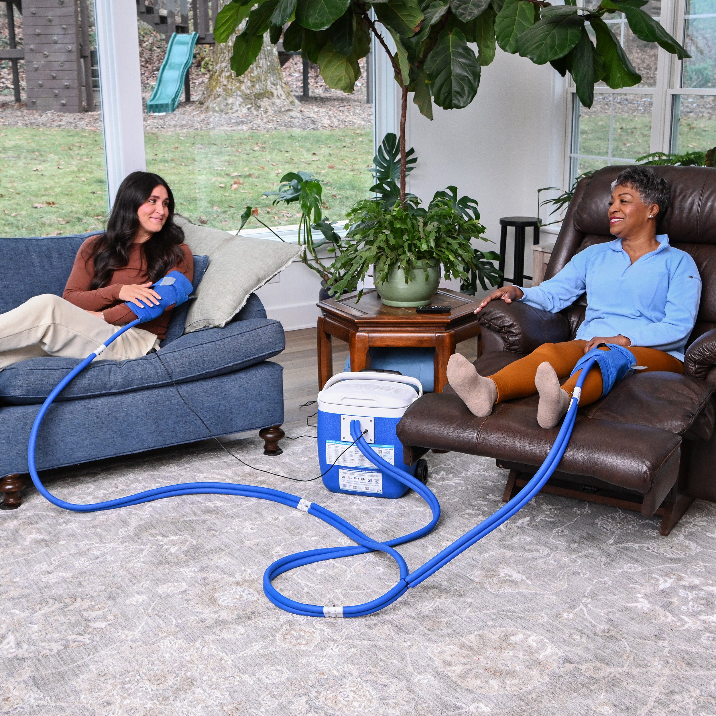 Two women sitting on a couch and chair with a blue hose connected to an active ice® cold therapy system in a living room.