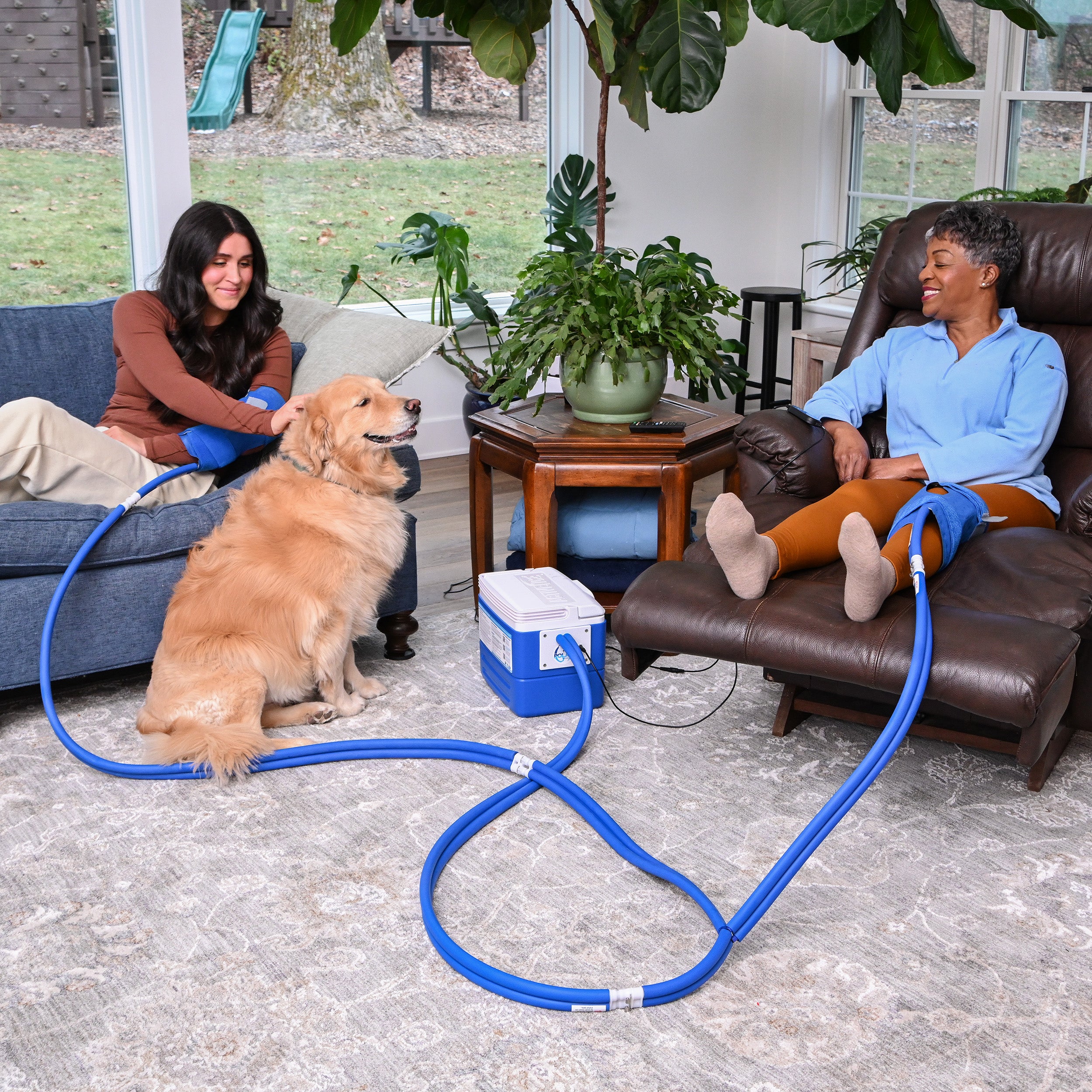 Two women sitting on a couch and chair and a gold retriever with a blue hose connected to an active ice® cold therapy system in a living room.