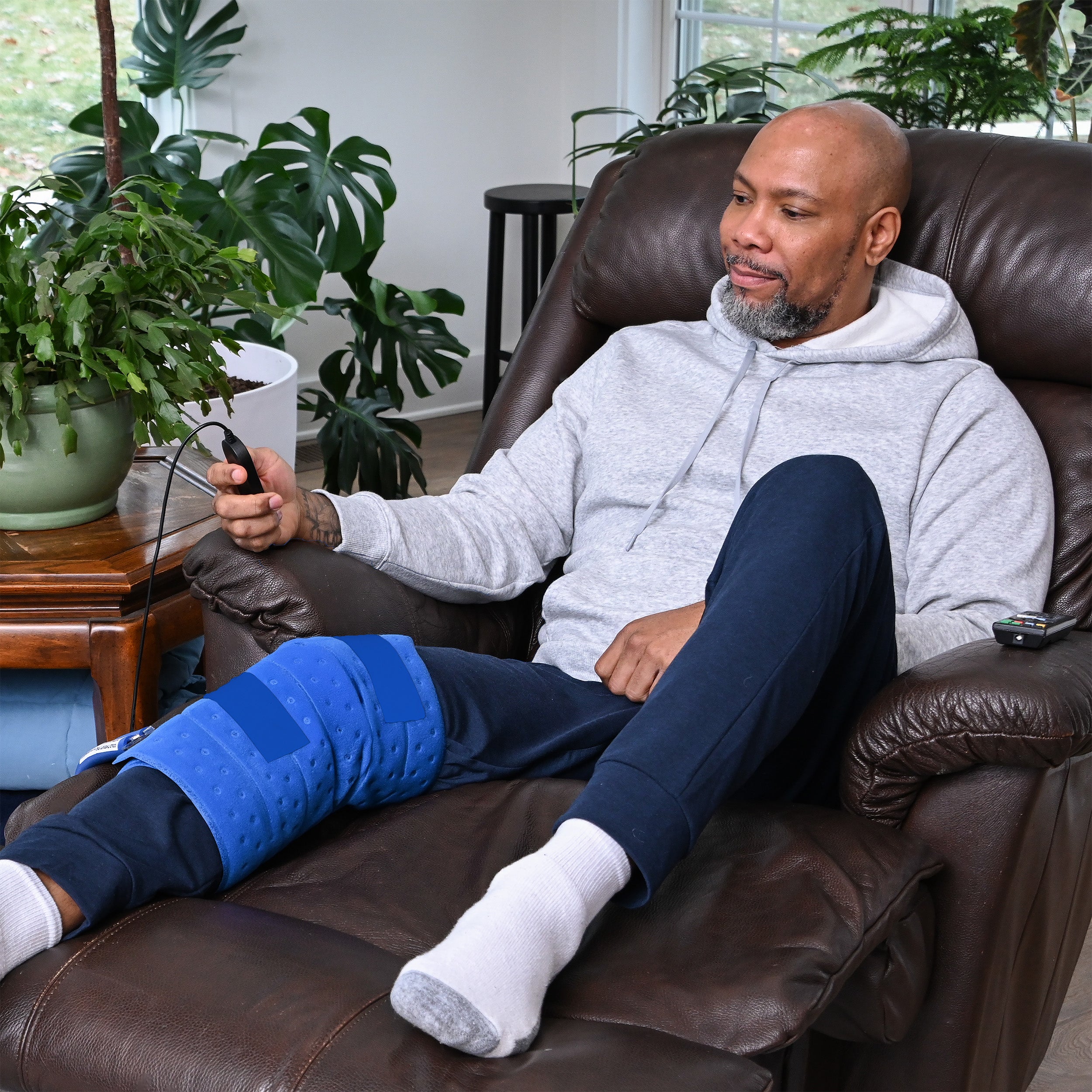 Man sitting on a brown leather chair in a living room with plants around with Full Circumference Knee Therapy Pad on knee
