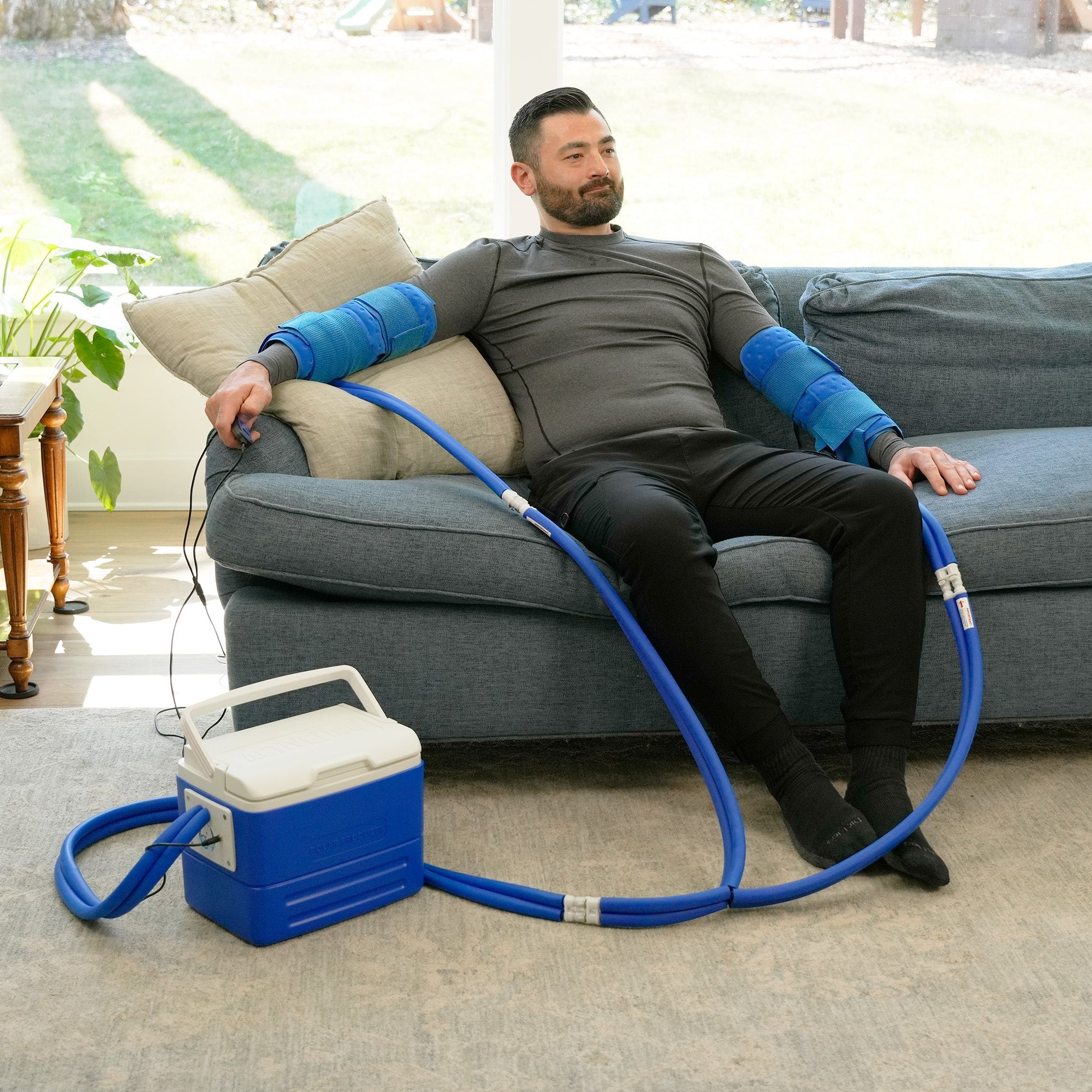 Man sitting on a couch using the Double Universal Rectangular Cold Therapy System on both of his arm in a living room setting