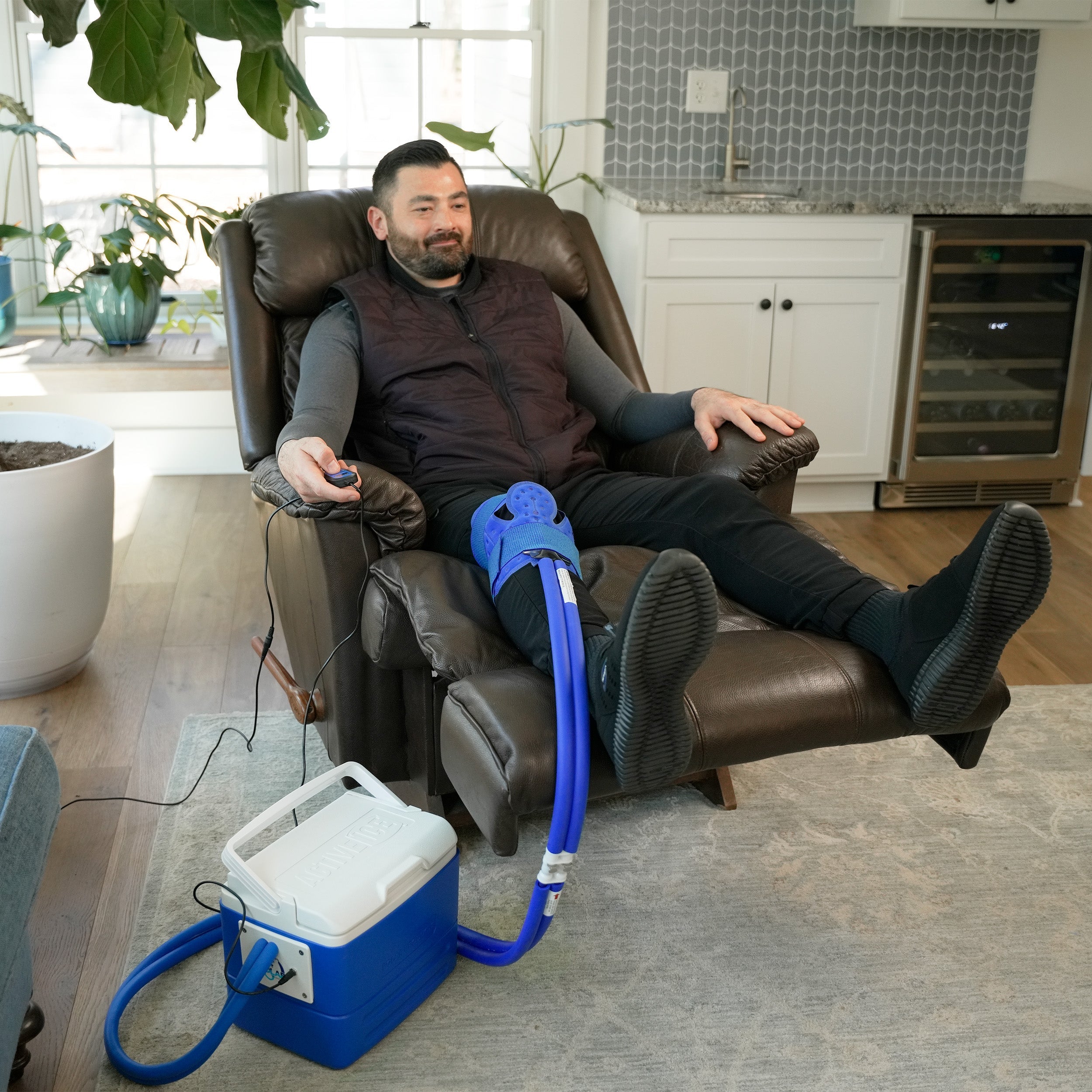 Man sitting in a recliner with a blue Universal Cold Therapy System on his knee, in a home setting.