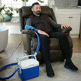 A man sits in a brown leather recliner in a home setting, wearing a blue Active Ice® Universal "U" Cold Therapy System on his elbow. A blue and white portable cooling device connected by tubing is placed on the floor beside him.