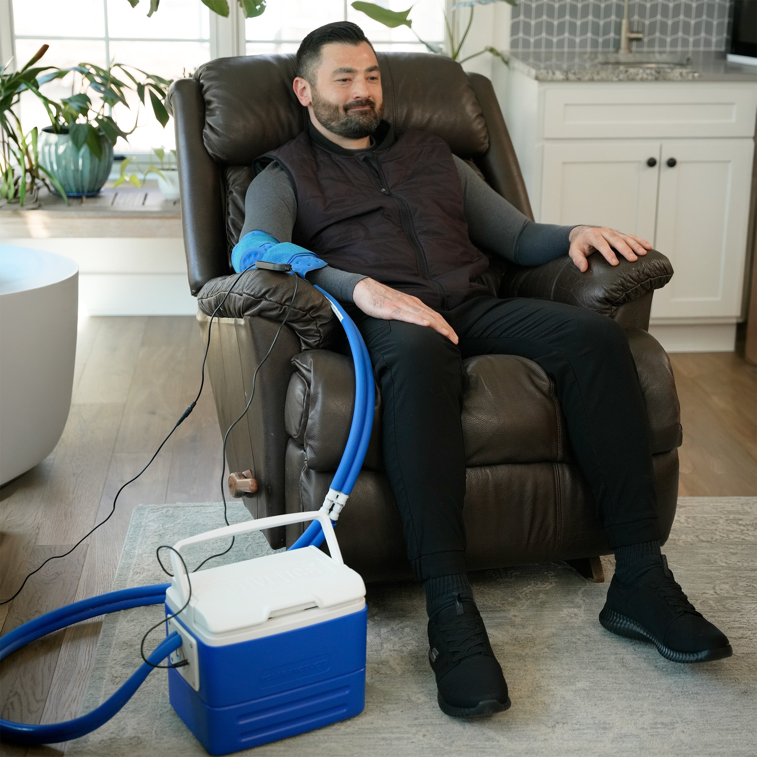 A man sits in a brown leather recliner in a home setting, wearing a blue Active Ice® Universal "U" Cold Therapy System on his elbow. A blue and white portable cooling device connected by tubing is placed on the floor beside him.