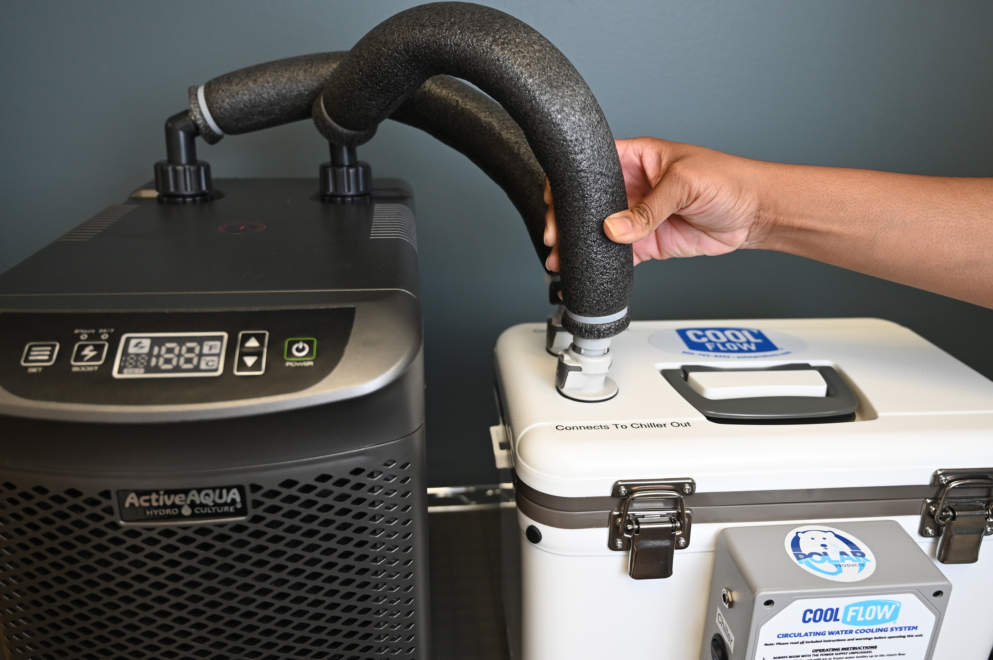 A person's hand connects a black foam insulated tube to a white cooler, next to an Arctic Chiller cooler system with a digital display screen.
