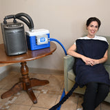 A woman sits in a chair wrapped in a dark navy circulating cooling blanket, connected by a blue hose to a 9-quart cooling reservoir and a gray Arctic Chiller on a wooden table. The cooler has a visible 'COOL Flow' label.