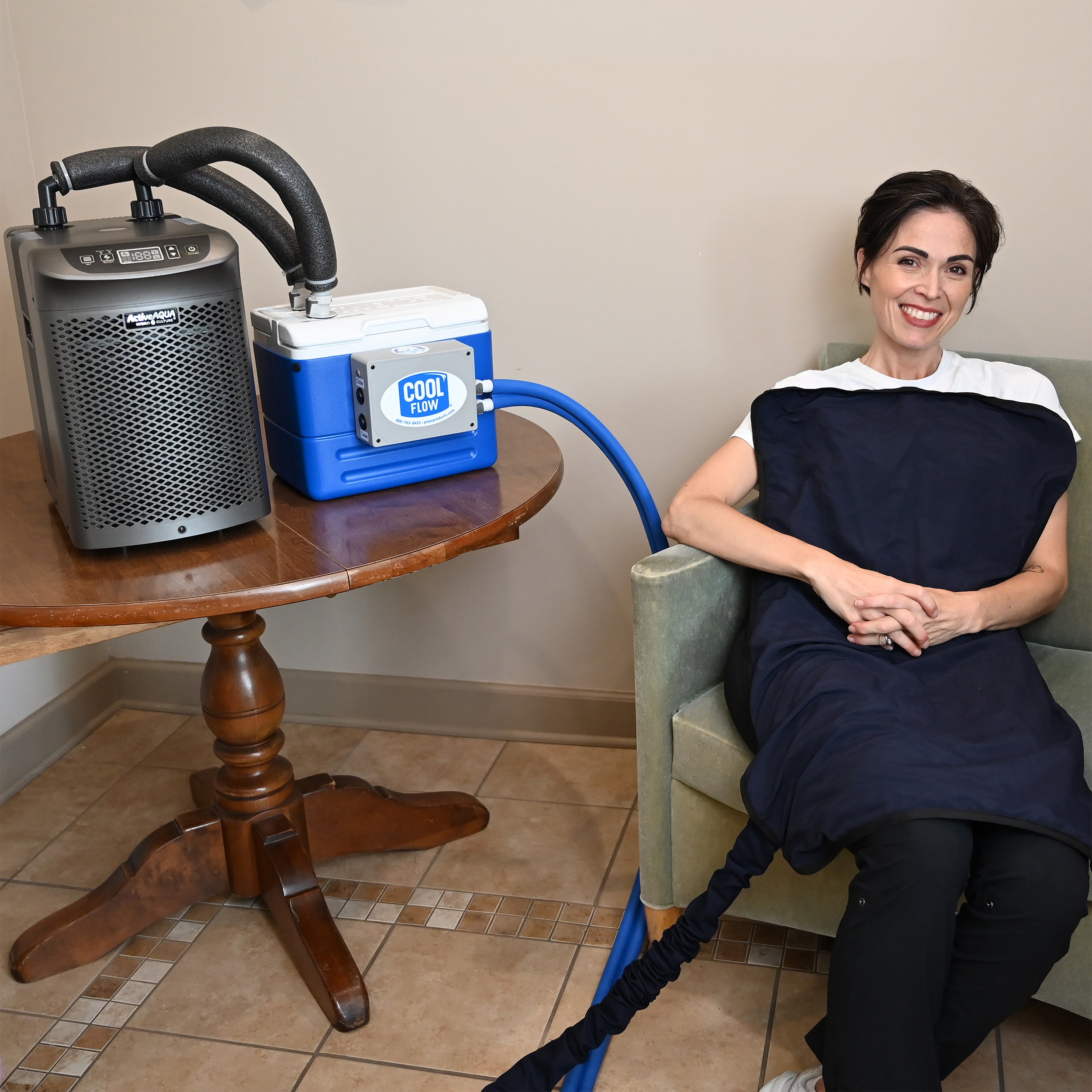 A woman sits in a chair wrapped in a dark navy circulating cooling blanket, connected by a blue hose to a 9-quart cooling reservoir and a gray Arctic Chiller on a wooden table. The cooler has a visible 'COOL Flow' label.