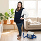 A woman stands in a bright living room wearing a dark navy Cool Flow® Adjustable Cooling vest in cotton over a white long-sleeve shirt and jeans, with a Cool Flow® device connected to her vest on the floor nearby.