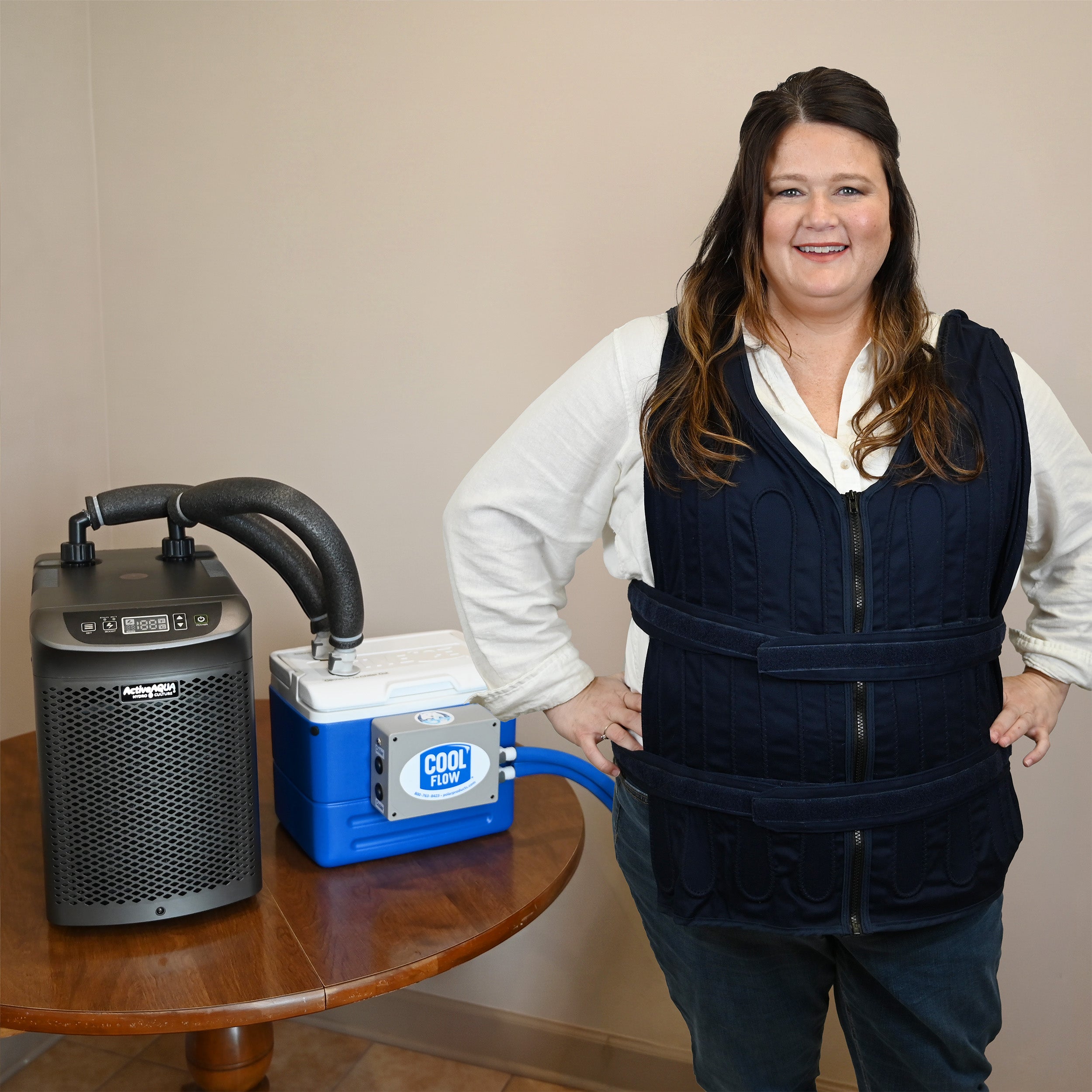 A woman stands indoors next to a portable 9-quart cooling reservoir attached to an Arctic Chiller on a wooden table, while wearing a dark navy adjustable circulating cooling vest.