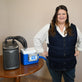 A woman stands indoors next to a portable 9-quart cooling reservoir attached to an Arctic Chiller on a wooden table, while wearing a dark navy adjustable circulating cooling vest.
