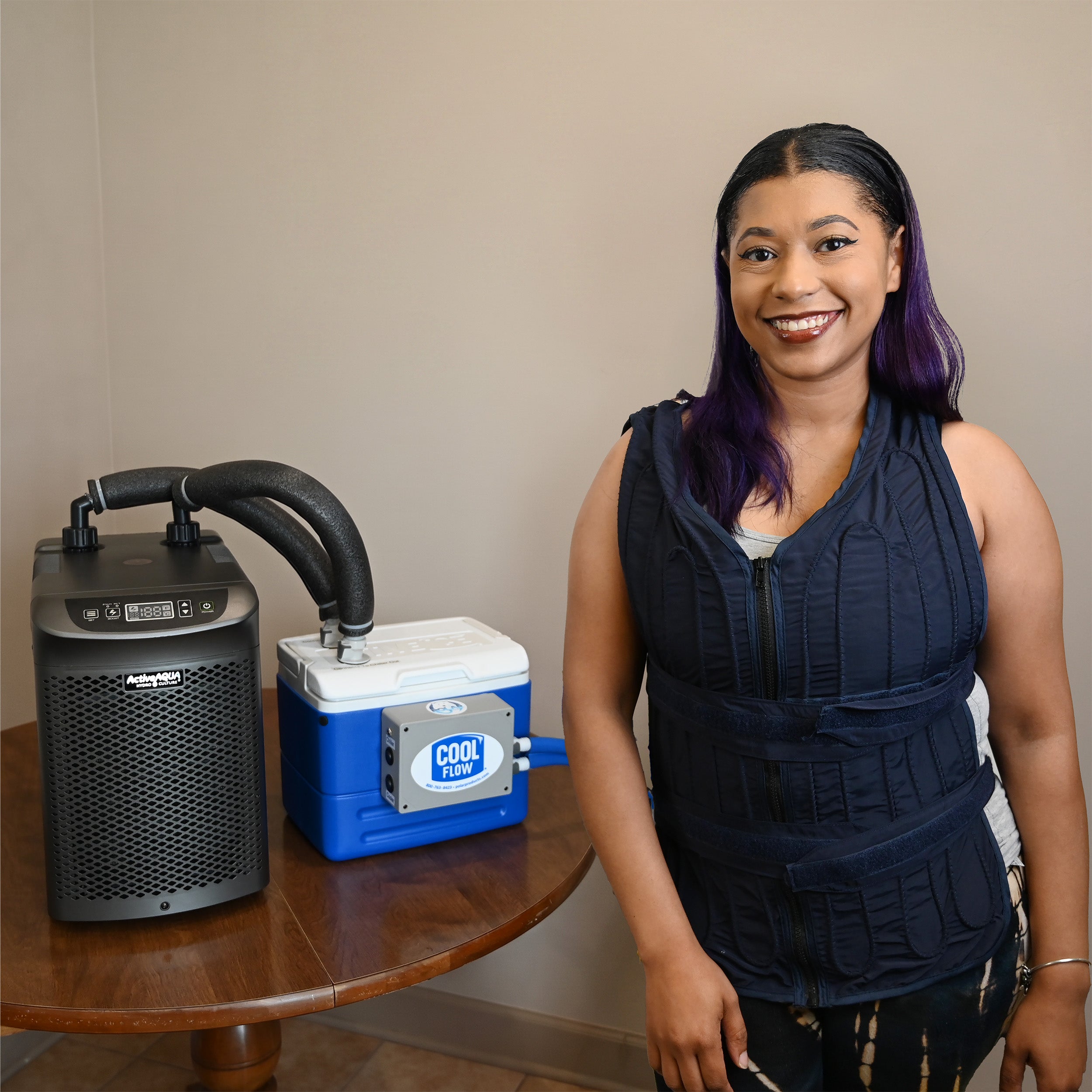 A woman stands indoors next to a portable 9-quart cooling reservoir attached to an Arctic Chiller on a wooden table, while wearing a dark navy adjustable circulating cooling vest.