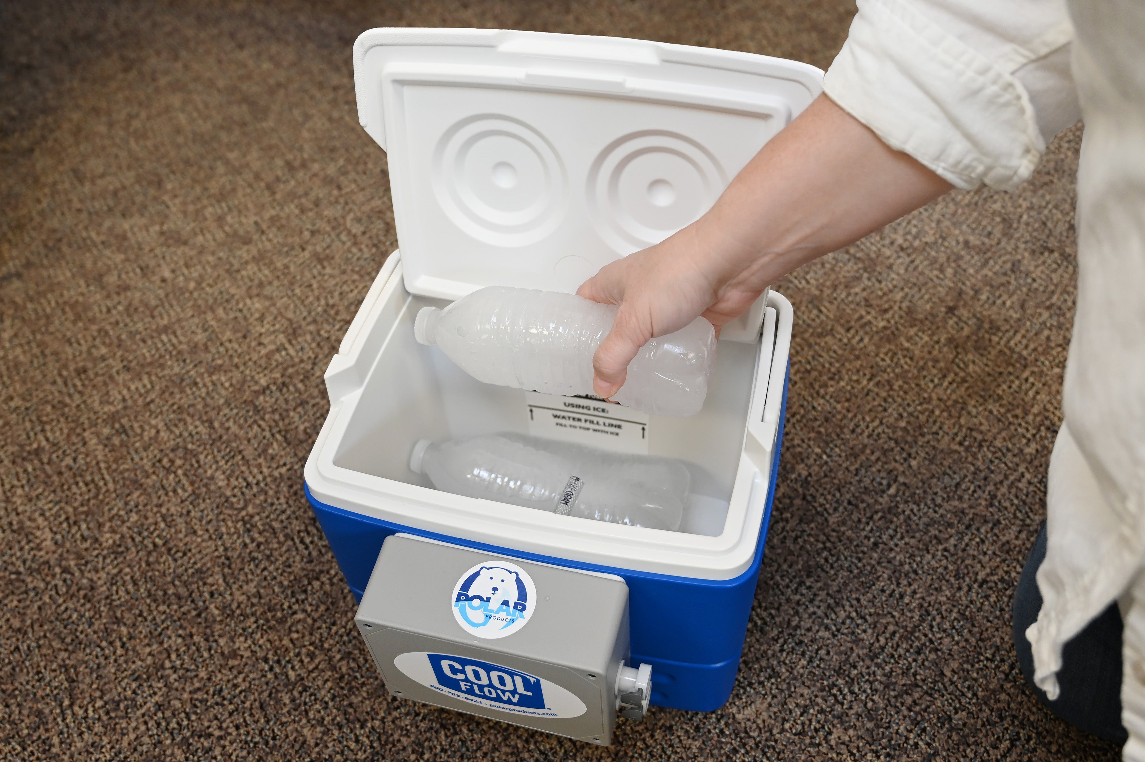 A person's hand placing a frozen plastic water bottle into an open blue and white Cool Flow® cooling reservoir with a gray COOLFLOW pump attached to the front, set on a brown carpeted floor.