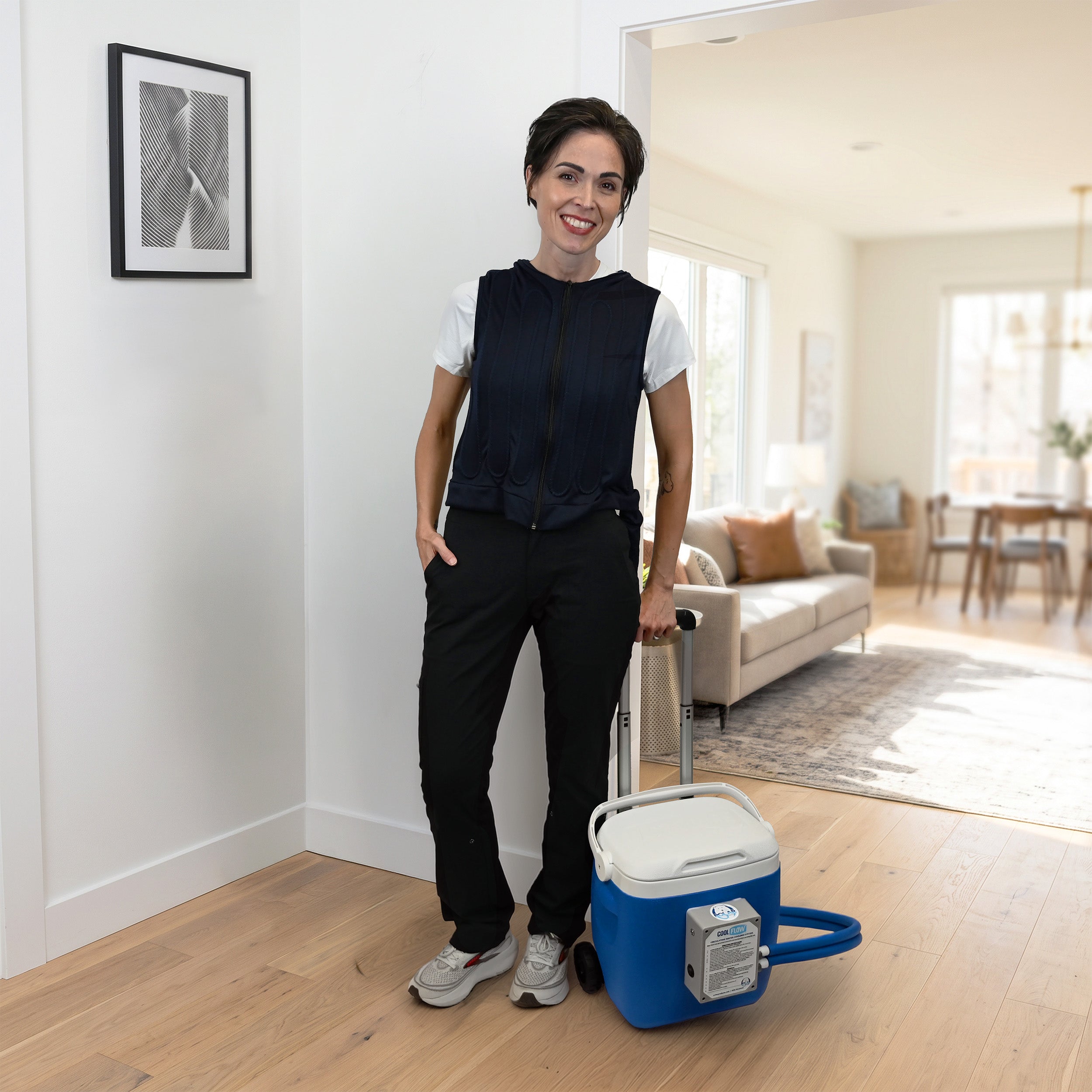 A woman stands in a bright, modern home interior next to a blue and white 19-quart cooling reservoir with wheels and retractable handle. She is wearing a navy fitted circulating cooling vest over a white t-shirt, black pants, and gray sneakers.