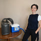 A woman stands indoors next to a portable 9-quart cooling reservoir attached to an Arctic Chiller on a wooden table, while wearing a dark navy fitted circulating cooling vest.
