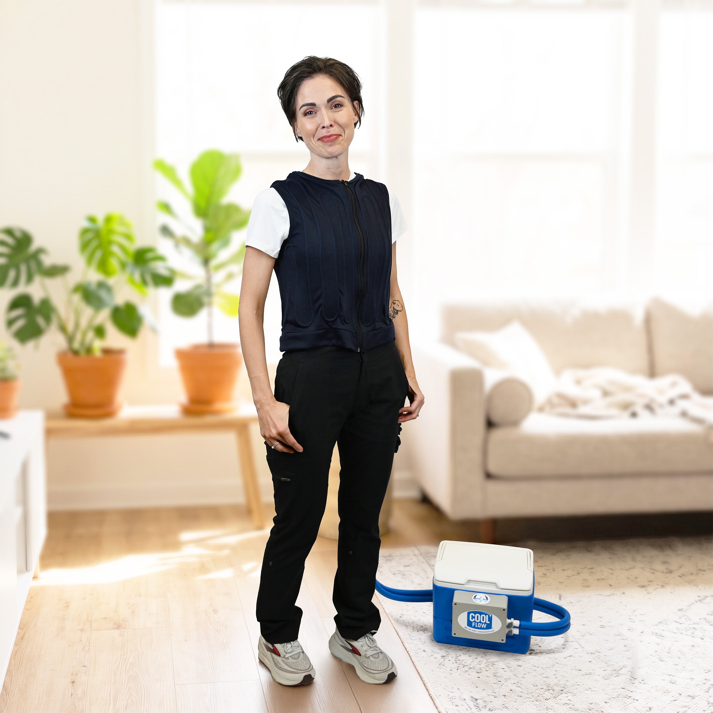A woman stands in a bright living room wearing a dark navy fitted circulating cooling vest over a white t-shirt, black cargo pants, and gray sneakers. A blue and white portable 9-quart cooling reservoir with a hose is on the floor beside her.