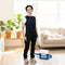 A woman stands in a bright living room wearing a dark navy fitted circulating cooling vest over a white t-shirt, black cargo pants, and gray sneakers. A blue and white portable 9-quart cooling reservoir with a hose is on the floor beside her.