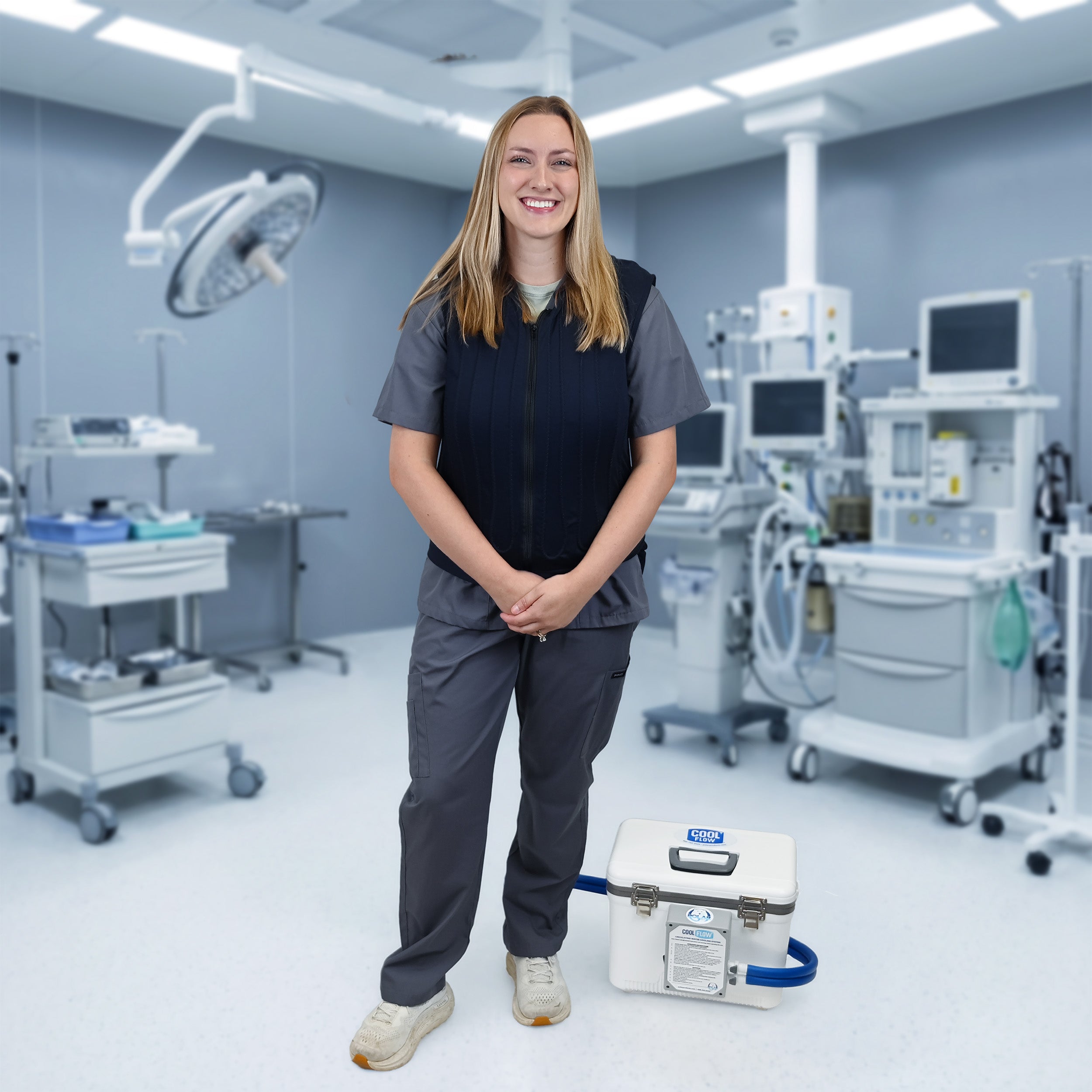A smiling female healthcare worker in dark gray scrubs and a navy fitted circulating cooling vest stands in a modern operating room, with a white 13-quart Cool Flow OR® cooling reservoir connected by a blue hose on the floor beside her.