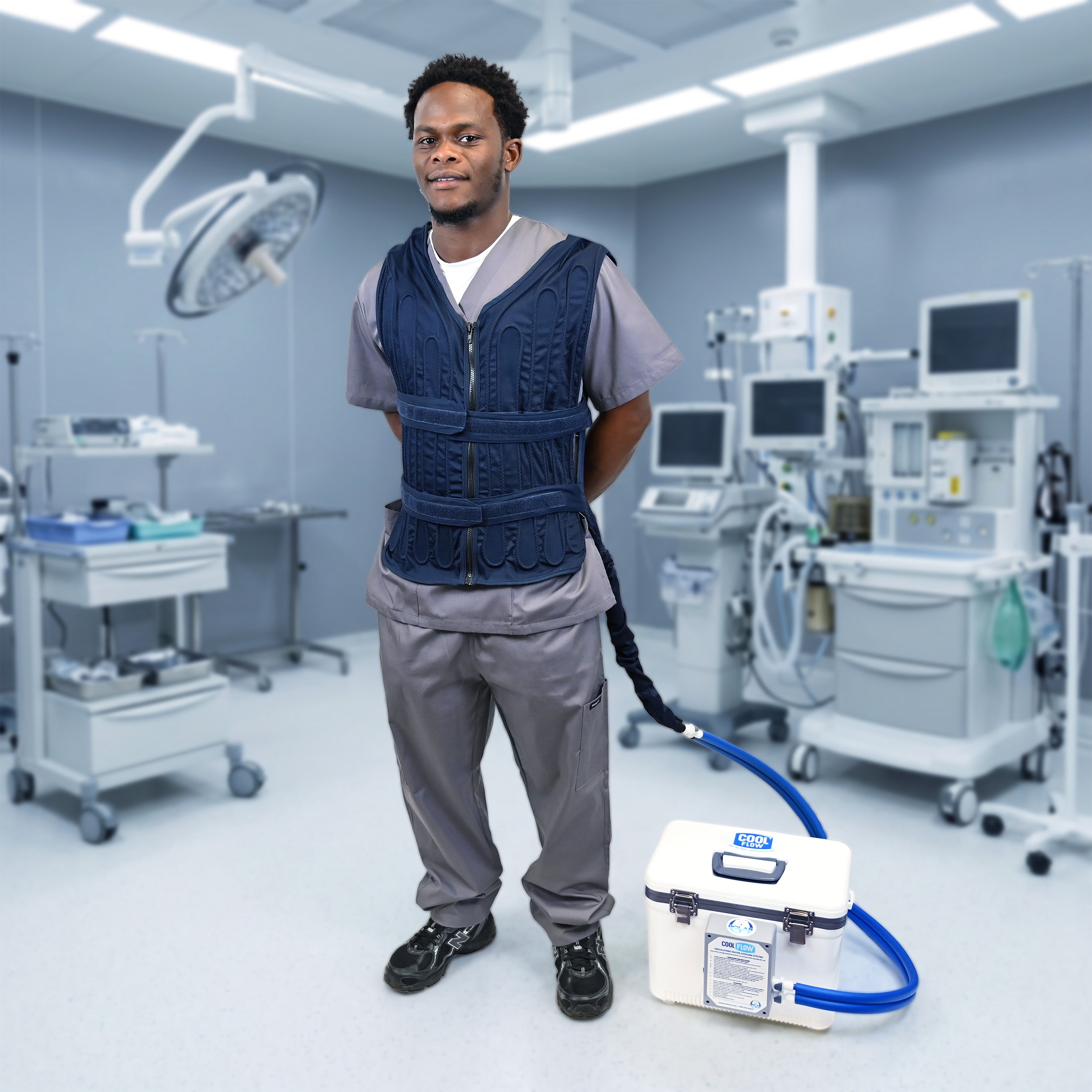 A man in medical scrubs stands in an operating room wearing a dark blue Cool Flow® circulating cooling vest, connected to a  13-quart cooling reservoir on the floor nearby.