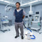 A man in medical scrubs stands in an operating room wearing a dark blue Cool Flow® circulating cooling vest, connected to a  13-quart cooling reservoir on the floor nearby.