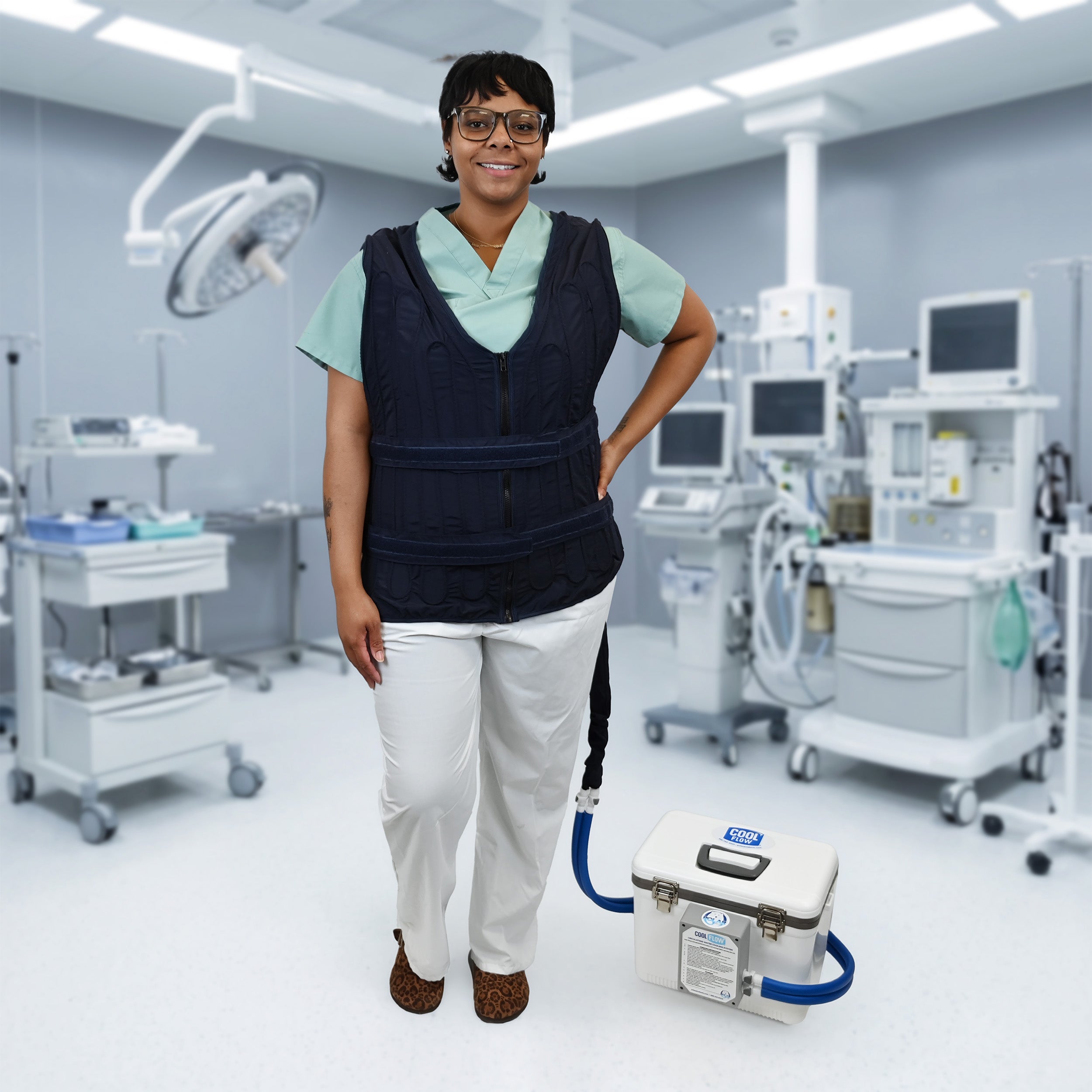 A woman in a medical room wearing a dark navy adjustable circulating cooling vest over a light green scrub top and white pants, standing next to a white 13-quart cooling reservoir with blue tubing.