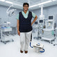 A woman in a medical room wearing a dark navy adjustable circulating cooling vest over a light green scrub top and white pants, standing next to a white 13-quart cooling reservoir with blue tubing.