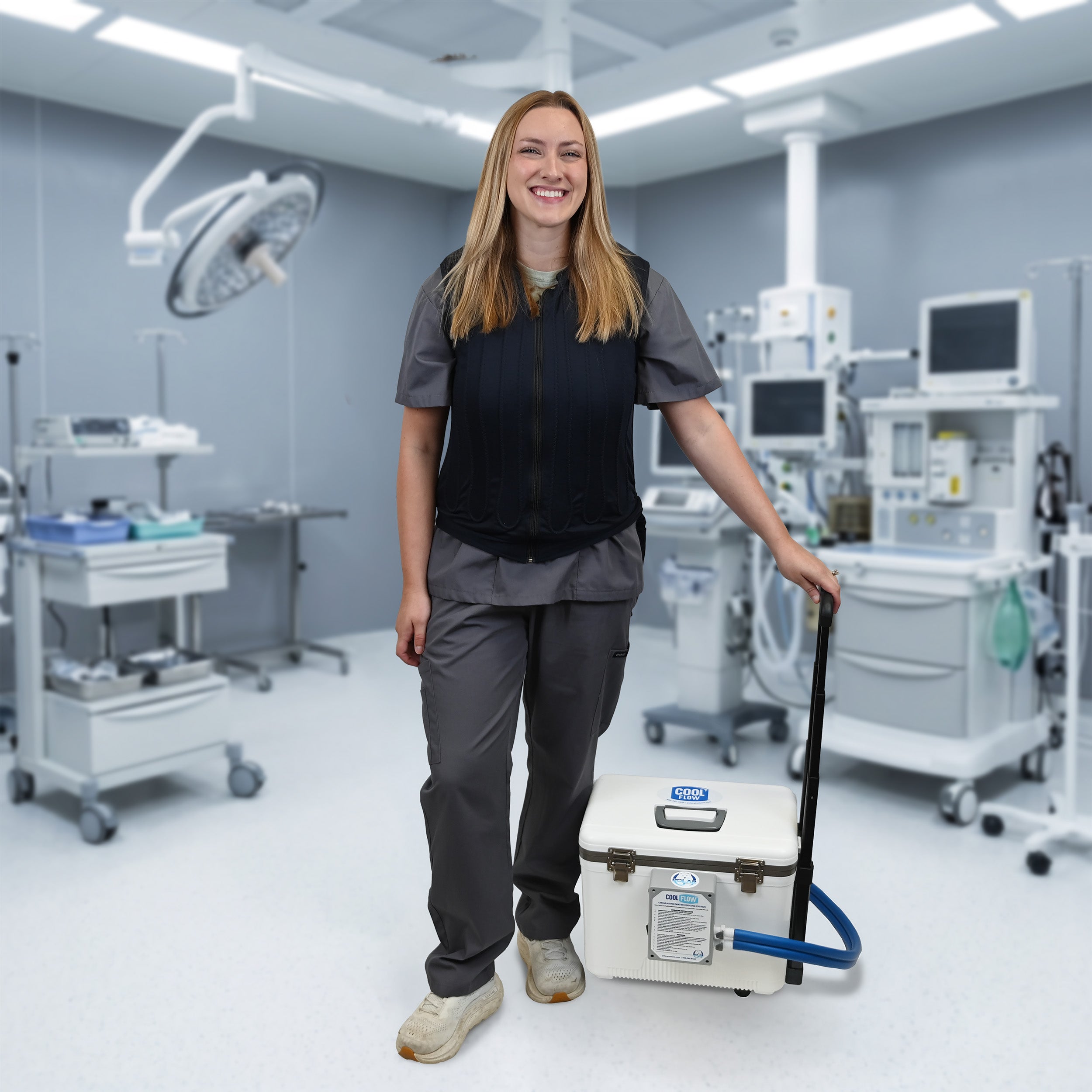 A woman in medical scrubs stands in a modern operating room holding a white portable 19-quart cooling reservoir with a blue hose and handle. She is wearing a navy blue fitted circulating cooling vest that is attached to the reservoir. 