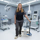 A woman in medical scrubs stands in a modern operating room holding a white portable 19-quart cooling reservoir with a blue hose and handle. She is wearing a navy blue fitted circulating cooling vest that is attached to the reservoir. 