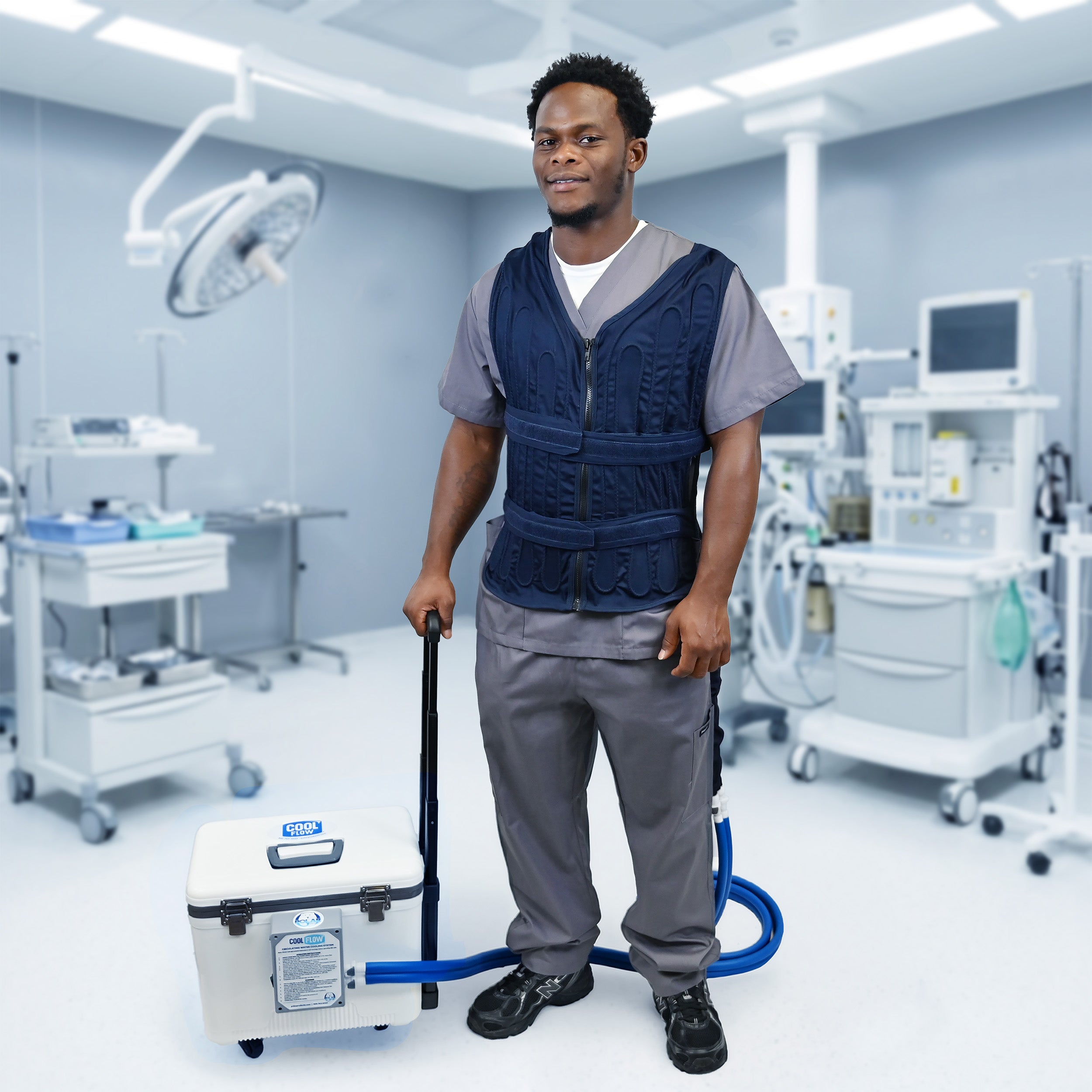 A man in medical scrubs stands in an operating room wearing a dark blue Cool Flow® circulating cooling vest, connected to a  19-quart cooling reservoir with a retractable handle and wheels on the floor nearby.