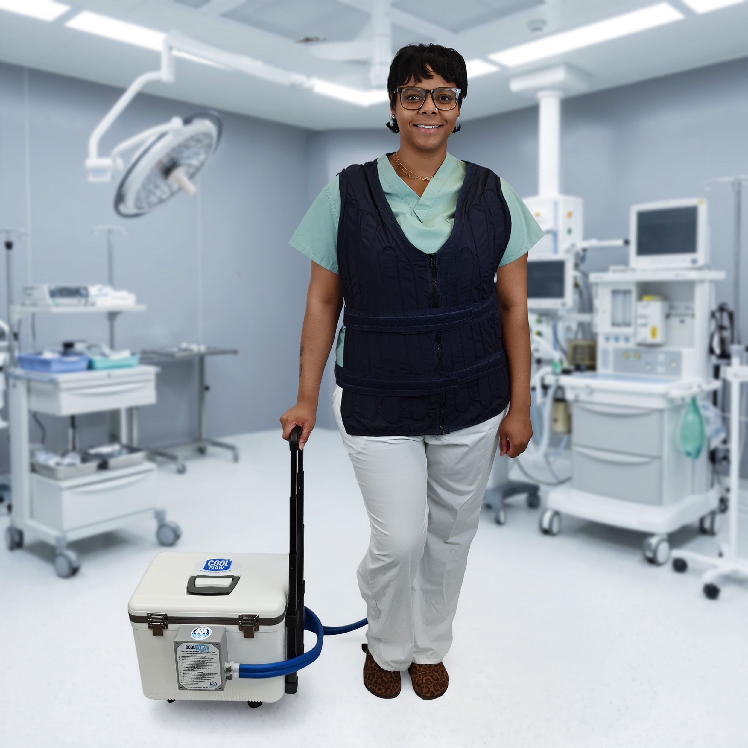 A woman in a medical room wearing a dark navy adjustable circulating cooling vest over a light green scrub top and white pants, standing next to a white 19-quart cooling reservoir with wheels, a retractable handle, and blue tubing.