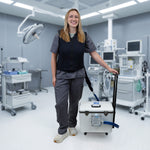 A smiling female healthcare worker in dark gray scrubs and a navy fitted circulating cooling vest stands in a modern operating room, with a white 30-quart Cool Flow OR® cooling reservoir with wheels and retractable handle connected by a blue hose on the floor beside her.