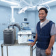 A man in medical scrubs stands beside a tiered cart holding two pieces of equipment: a dark gray Arctic Chiller with a hose and a white 13-quart cooling reservoir, set against a blurred operating room background. The cooling reservoir is attached to the adjustable circulating cooling vest that the man is wearing.
