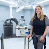 A woman in medical scrubs stands beside a metal cart holding an Arctic Chiller and 13-quart cooling reservoir in a bright operating room. She's attached to the reservoir with the fitted circulating cooling vest. The background shows surgical lights and medical equipment.